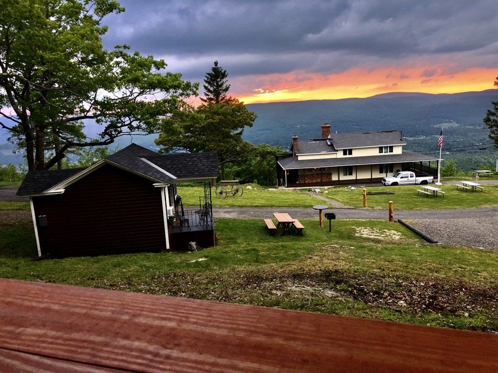 A sunset illuminates a mountainous landscape with a small wooden cabin in the foreground and a larger building nearby.