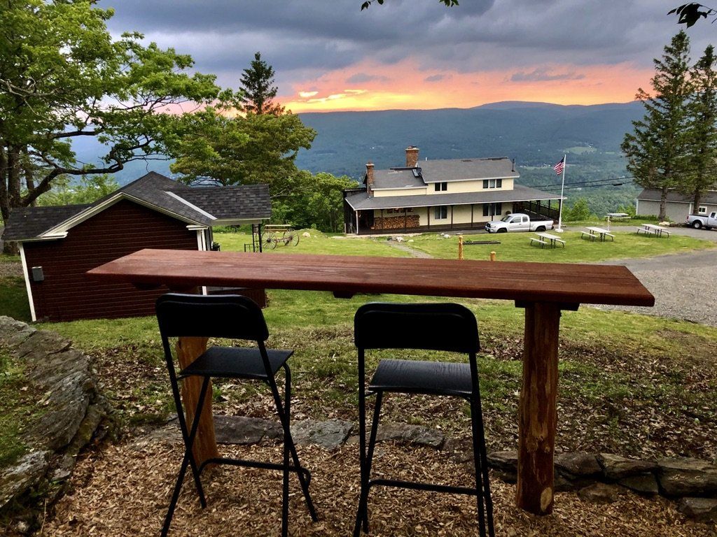 Two black bar stools stand before a wooden outdoor bar overlooking a sunset-lit valley with a house and trees.