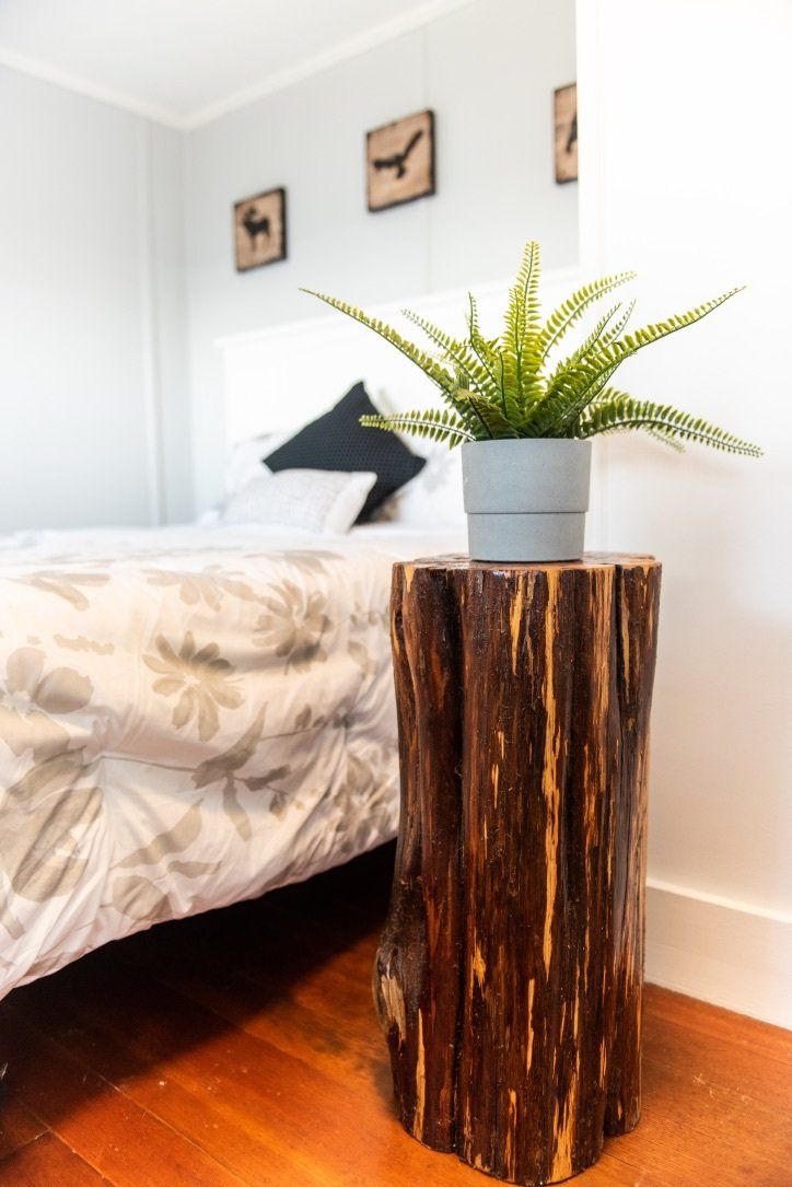 A green potted fern sits on a rustic wooden log side table in a bedroom next to a bed with patterned bedding.