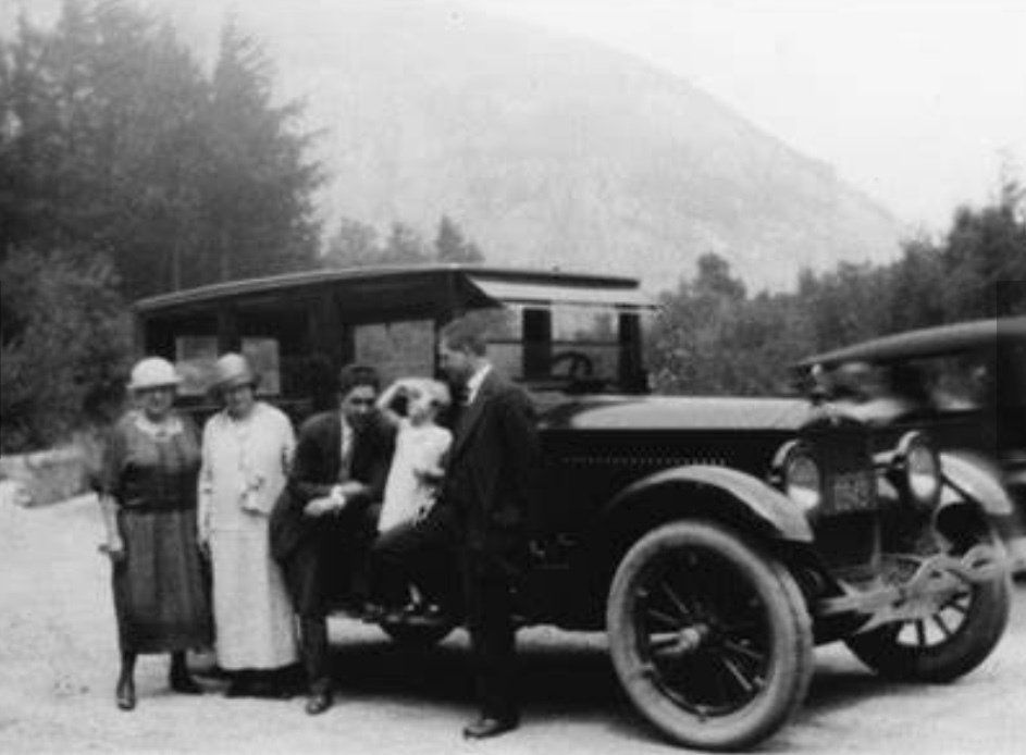 A black and white photo of a group standing beside a vintage car in a mountainous, wooded landscape.