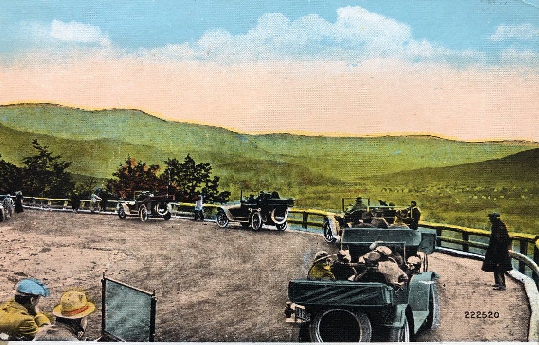 A scenic, hand-tinted postcard showing vintage cars parked at a mountain lookout with people enjoying the panoramic view.