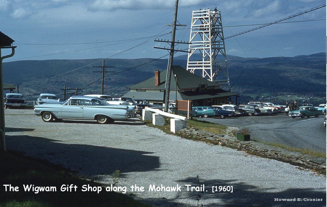A 1960 view of The Wigwam Gift Shop, featuring a parked light-blue car, a tall observation tower, and mountain views.