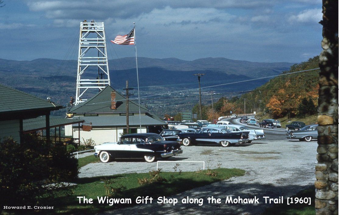 A 1960 view of The Wigwam Gift Shop on the Mohawk Trail, featuring a white metal tower, cars in a lot, and mountains.
