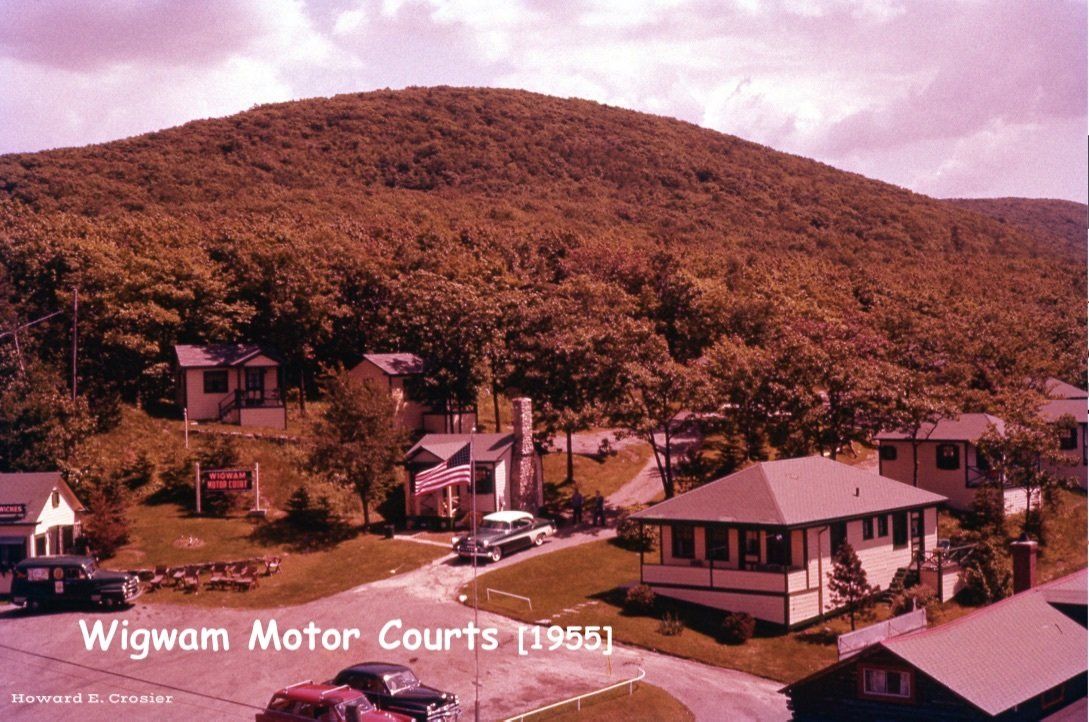 A 1955 photo of Wigwam Motor Courts, featuring small cottages near a wooded hillside with an American flag flying.