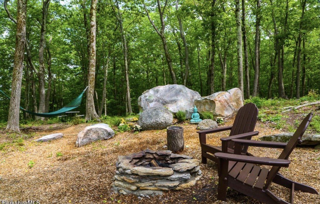 A fire pit and two brown Adirondack chairs in a clearing surrounded by trees, with a hammock and large rocks in the back.