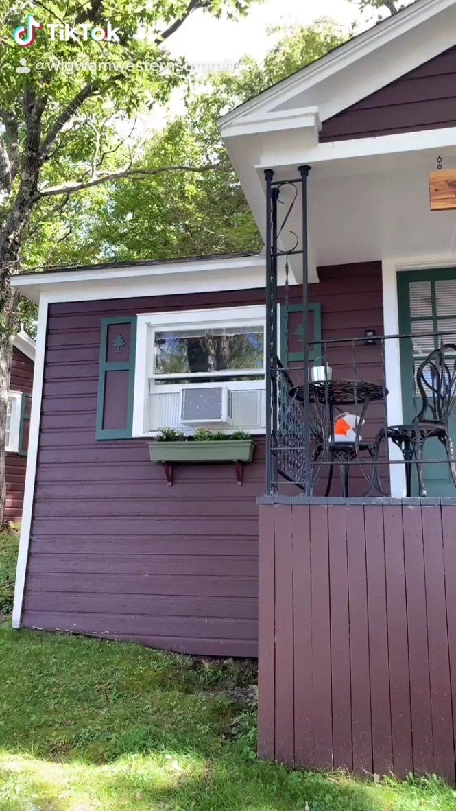 A small, dark purple house with a front porch, a green window box, and an air conditioning unit in the window.