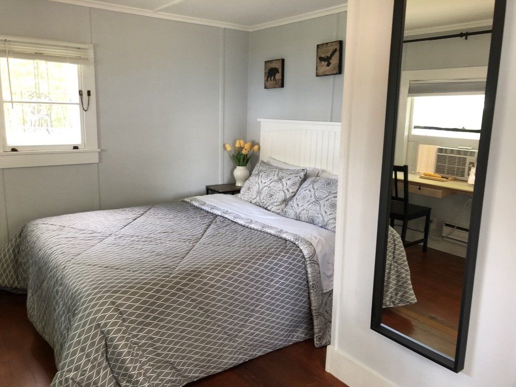 A bedroom with a bed featuring a gray patterned quilt, white headboard, small nightstand, and a full-length wall mirror.
