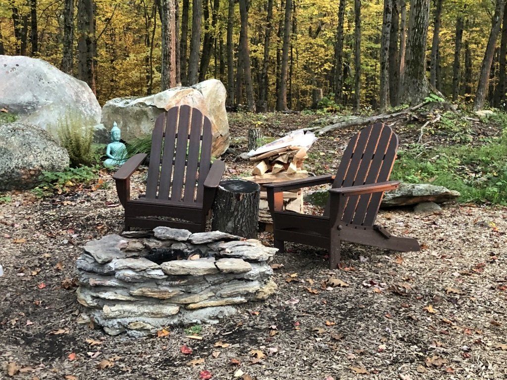 Two dark wood Adirondack chairs face a stone fire pit in a wooded area with fallen leaves.