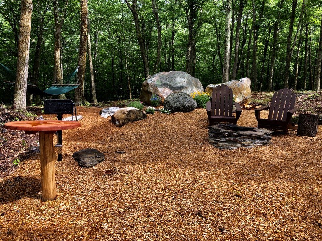A rustic outdoor seating area with two wooden chairs, a fire pit, and a small table on wood mulch in a shaded forest.