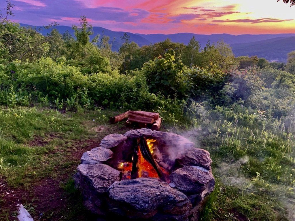 A stone fire pit with a crackling fire at dusk, set against a vibrant purple and orange sunset over rolling green hills.