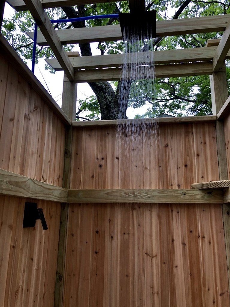 An outdoor cedar shower stall with a large square rain shower head actively flowing water.