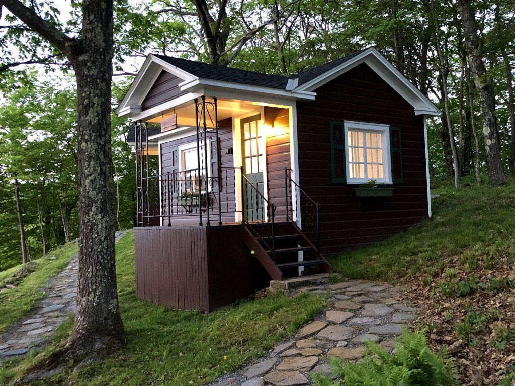 A small, dark brown cabin with a lit porch and stone walkway, nestled in a lush, green wooded area.