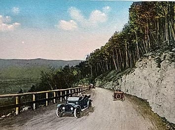 A vintage color postcard of two early automobiles driving along a winding mountain road with a wooden guardrail and forest.