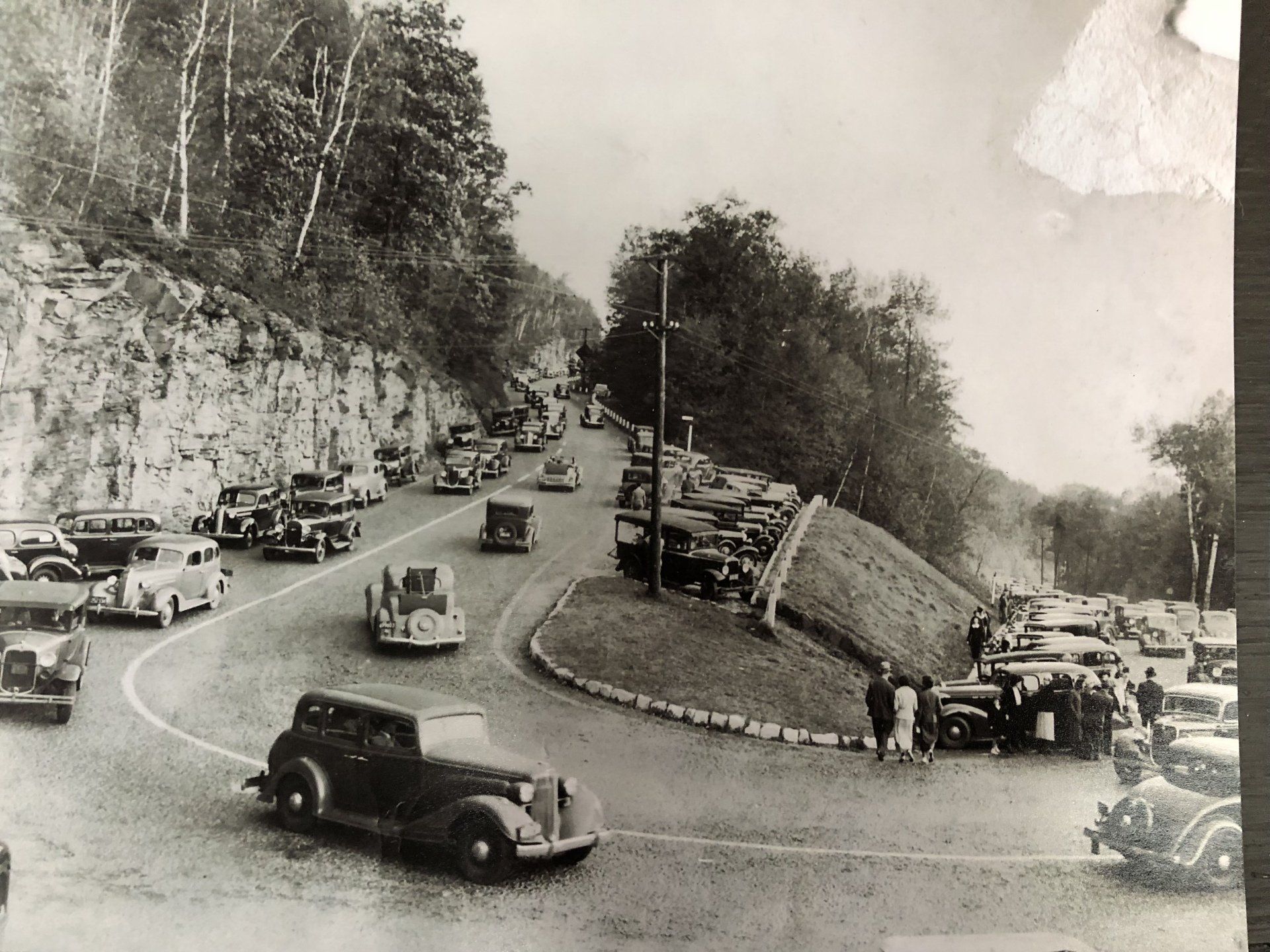 A vintage, black-and-white photograph shows a line of cars navigating a curved mountain road with trees and a rock wall.