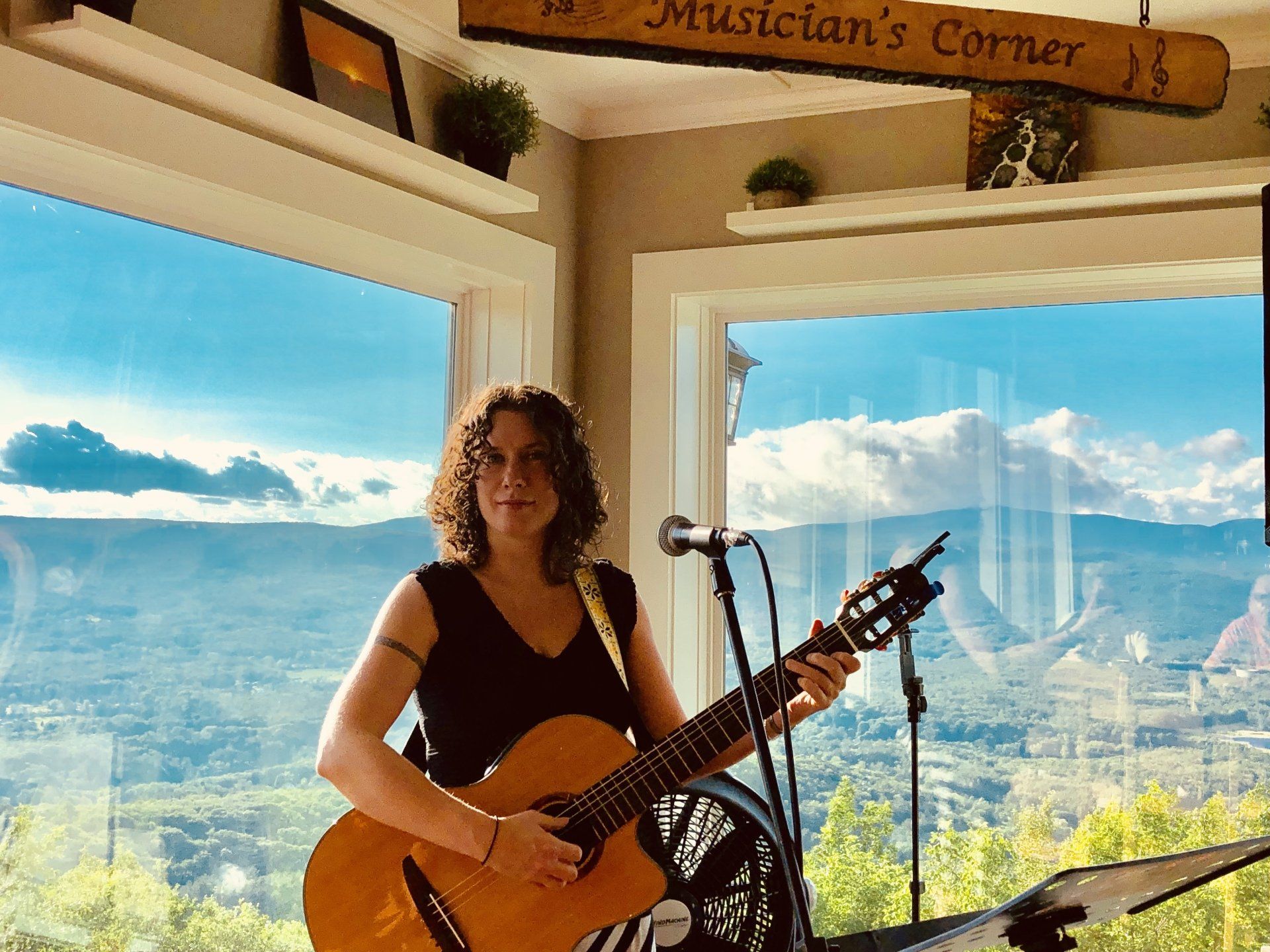 A person plays an acoustic guitar in a room with large windows overlooking a scenic mountain view, under a Musician's Corner sign.
