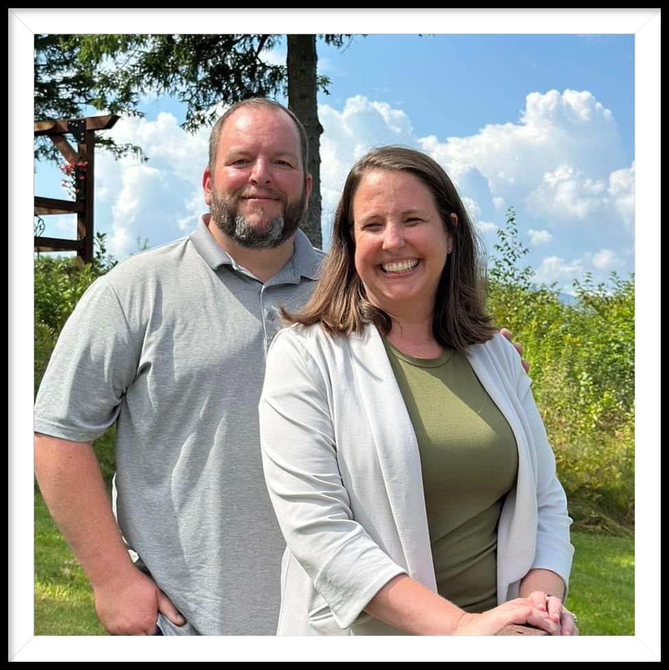 Wigwam Owners - Kelly and Alan smiling outdoors in a grassy area with trees and a blue sky in the background.
