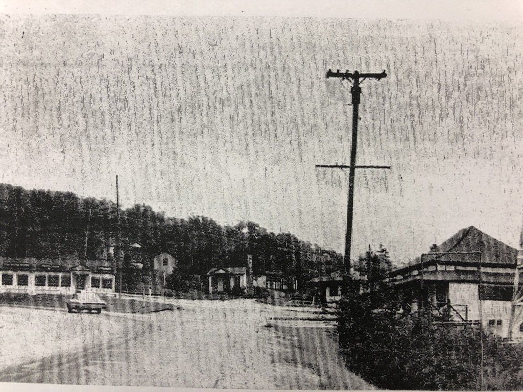 A black-and-white view of a dusty, rural road passing by small, light-colored buildings under a dense treeline.