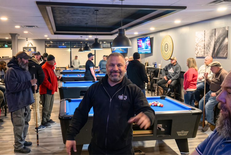 Man in black hoodie smiling in a busy pool hall with people around blue pool tables