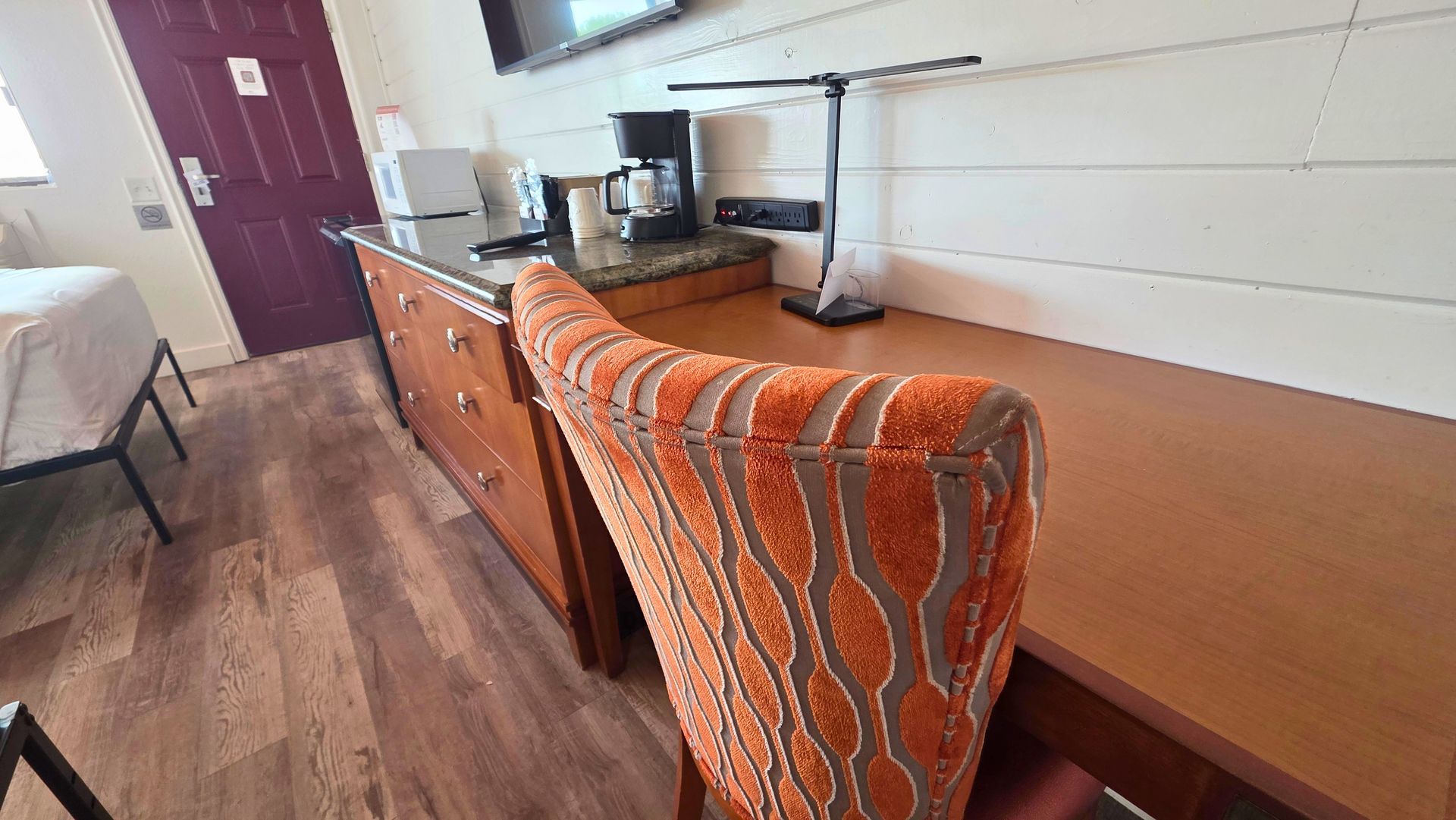 Dorm room desk with orange patterned chair, wood dresser, and TV on a countertop.