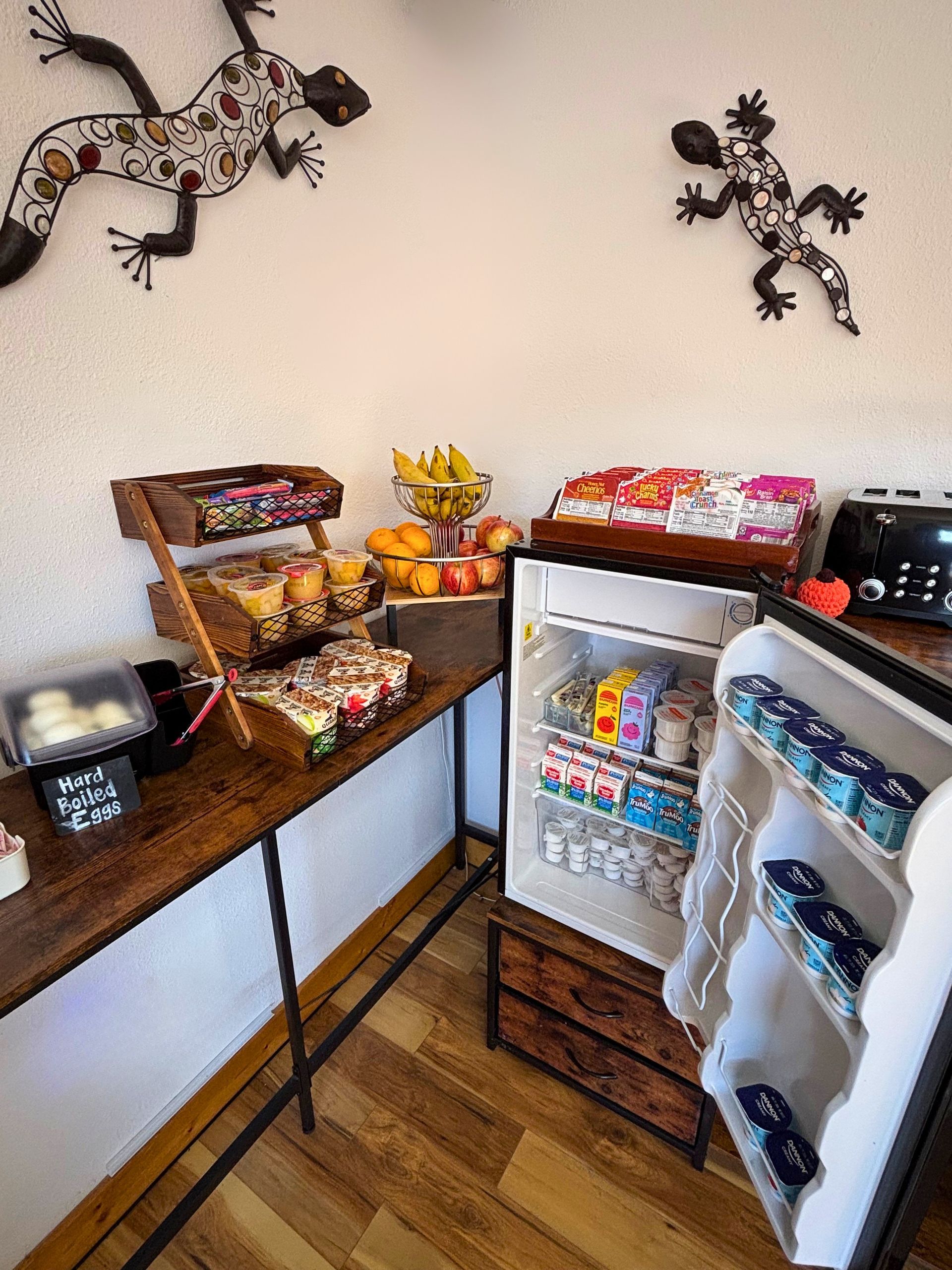 Small home drink station with mini fridge, snacks, and trays on a wooden floor and countertop.