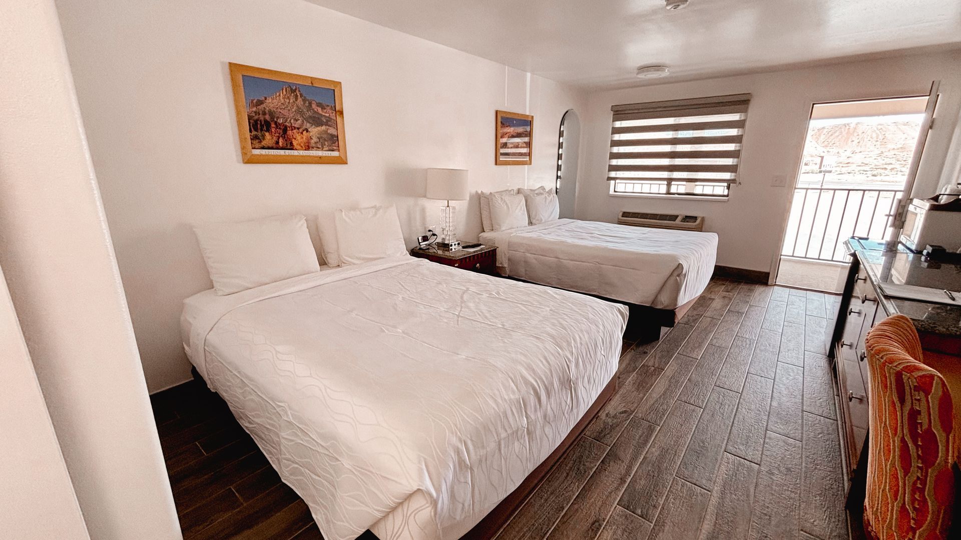 Bright hotel room with two white beds, wooden floor, and window blinds.