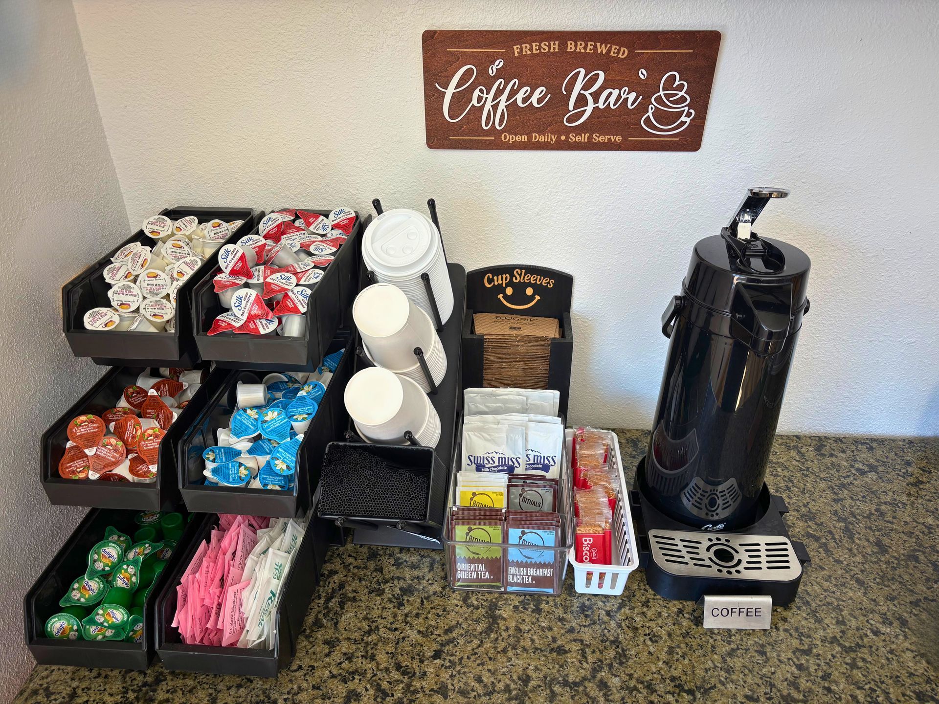 Coffee station with labeled hot beverage pods, cups, and a black coffee dispenser on a counter