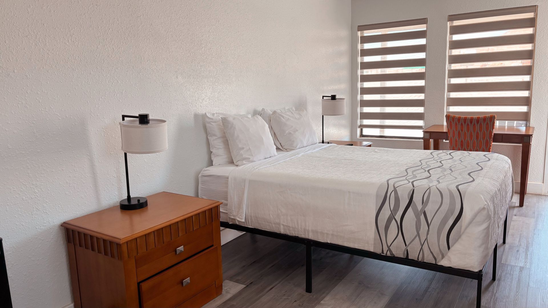 Bright bedroom with a white bed, wooden nightstand, striped blinds, and a window chair.