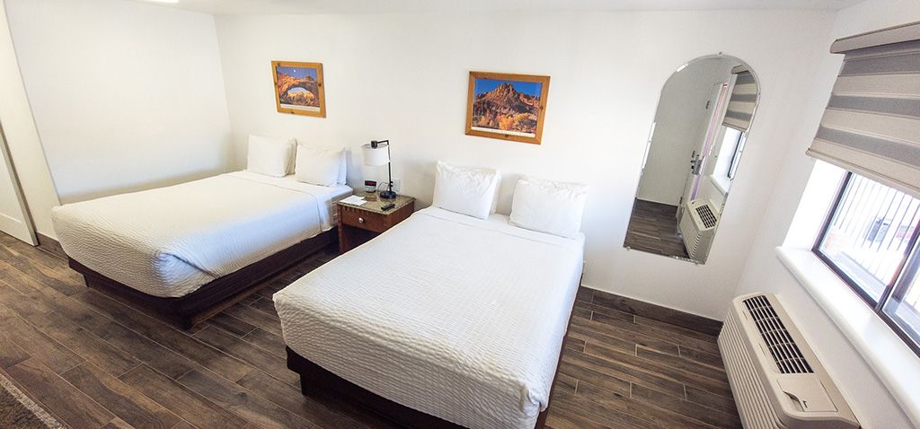 Two white beds in a simple hotel room with framed art, a mirror, and a window with a radiator under it
