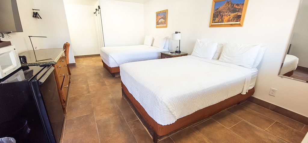 Small hotel room with two white beds, wooden floor, desk, and framed wall art