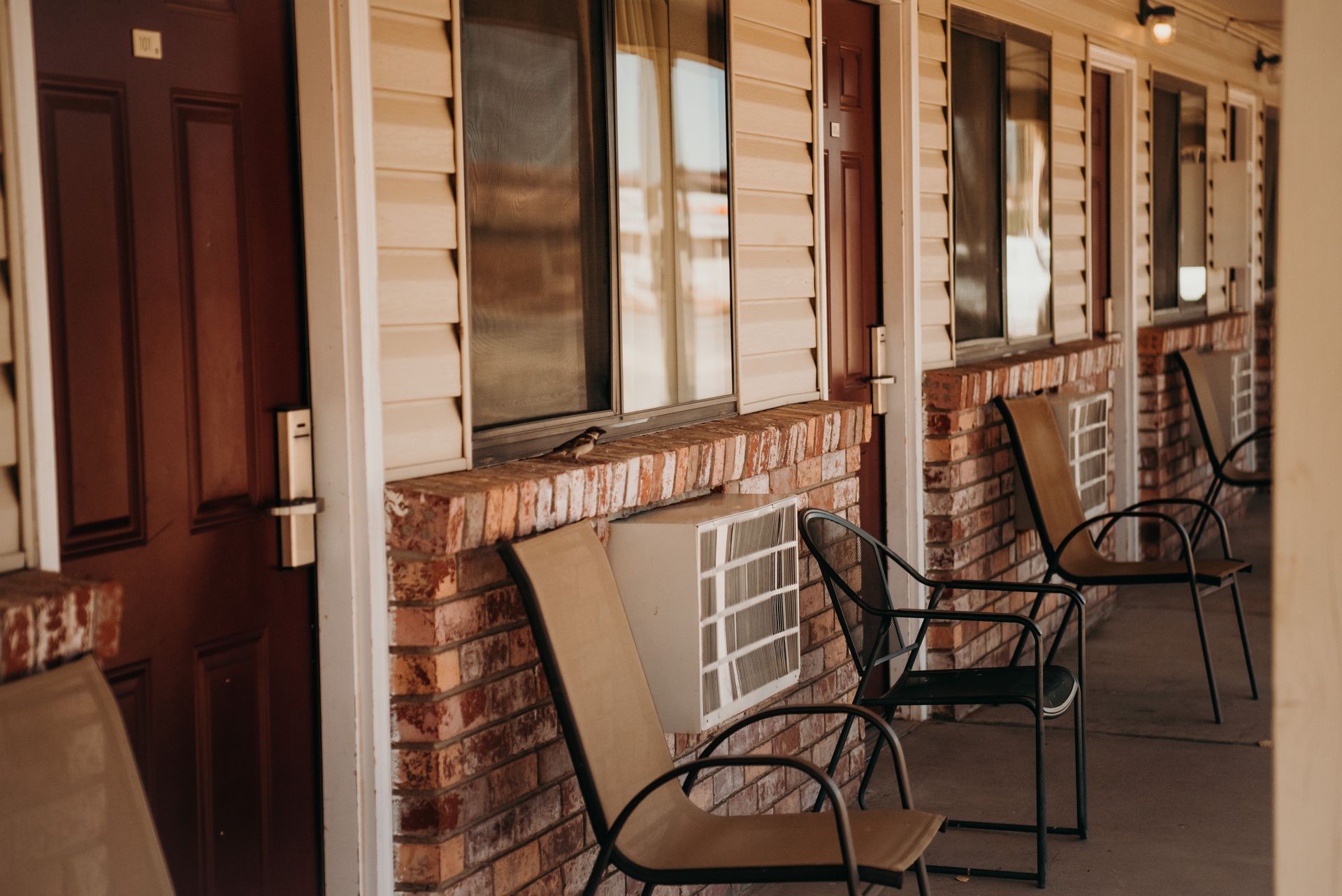 Porch with brick columns, metal chairs, and a wooden door on a sunny building exterior