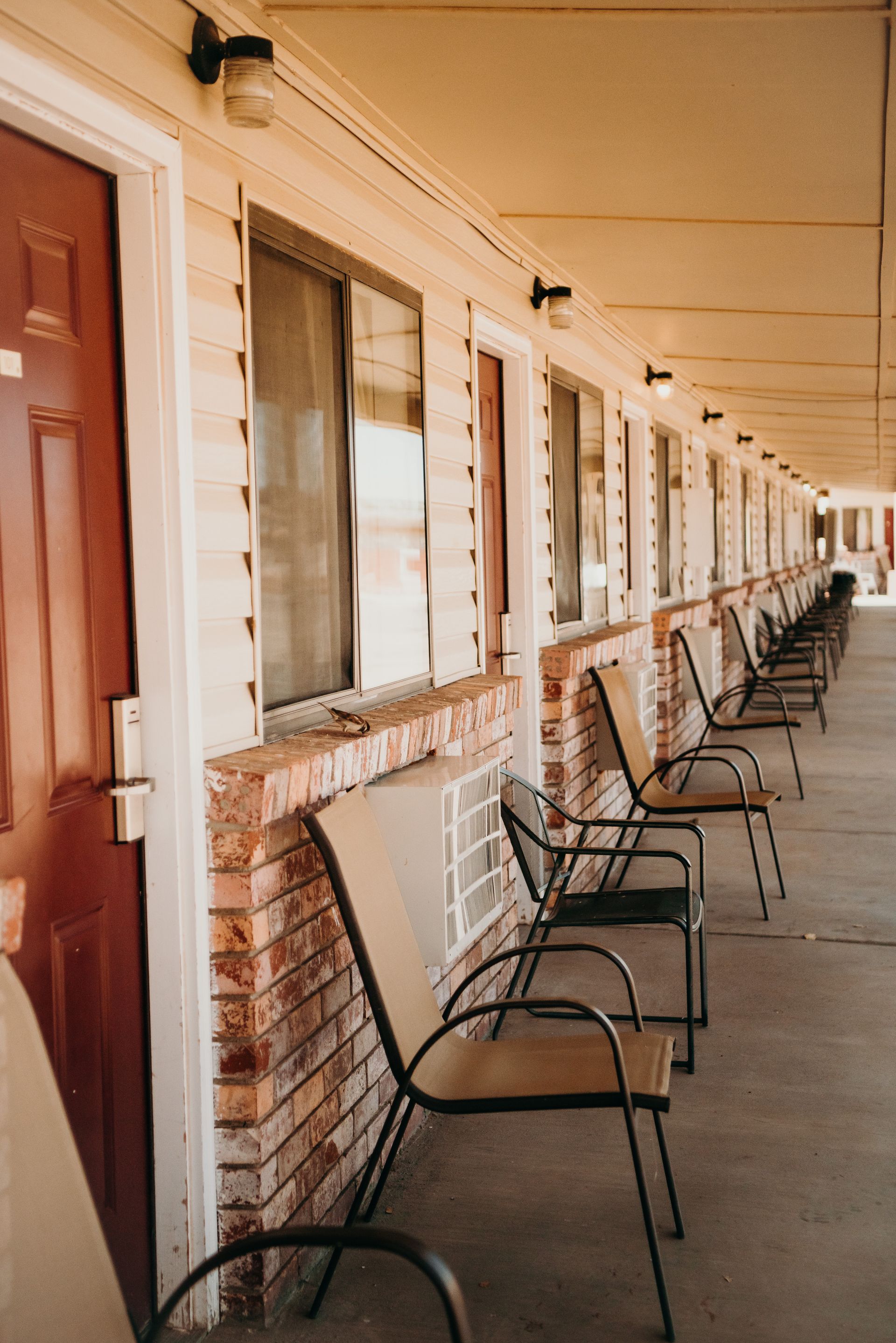 Exterior motel walkway with doors, windows, and black metal chairs along a covered corridor