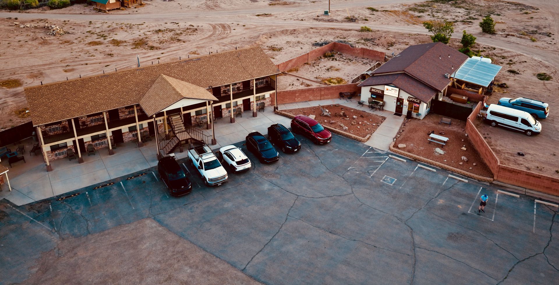 Aerial view of a desert motel parking lot with several cars, beige buildings, and a blue pickup truck.