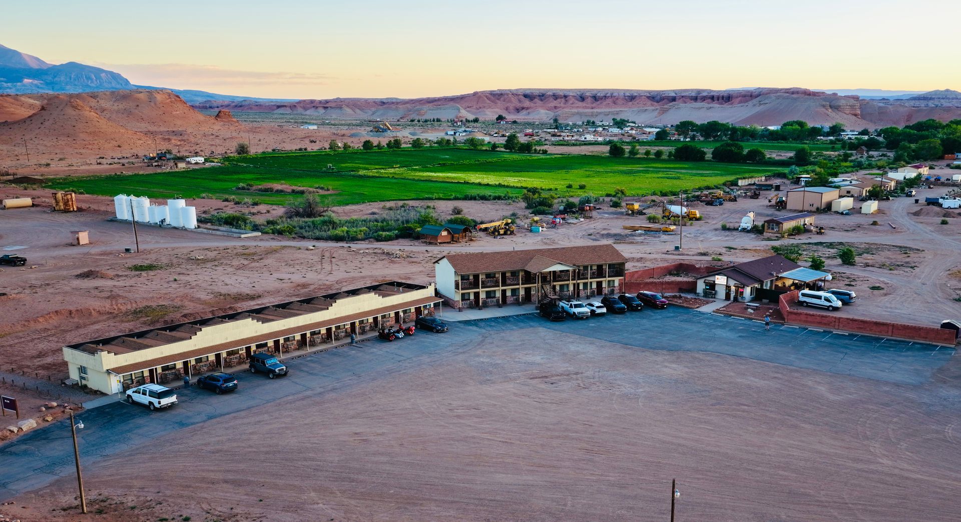 Desert resort with pools and buildings beside a green oasis at sunset, backed by rocky hills