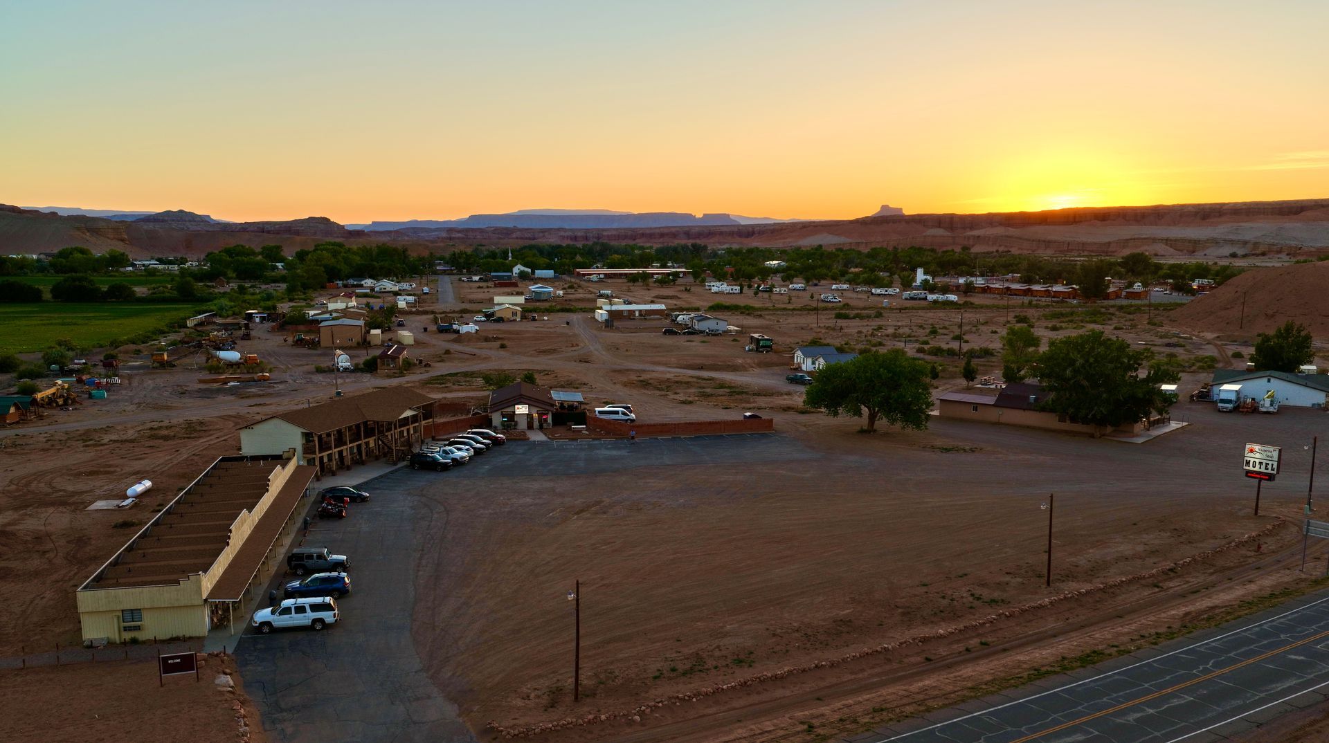 Sunset over a rural settlement with scattered homes, dirt roads, and parked cars.