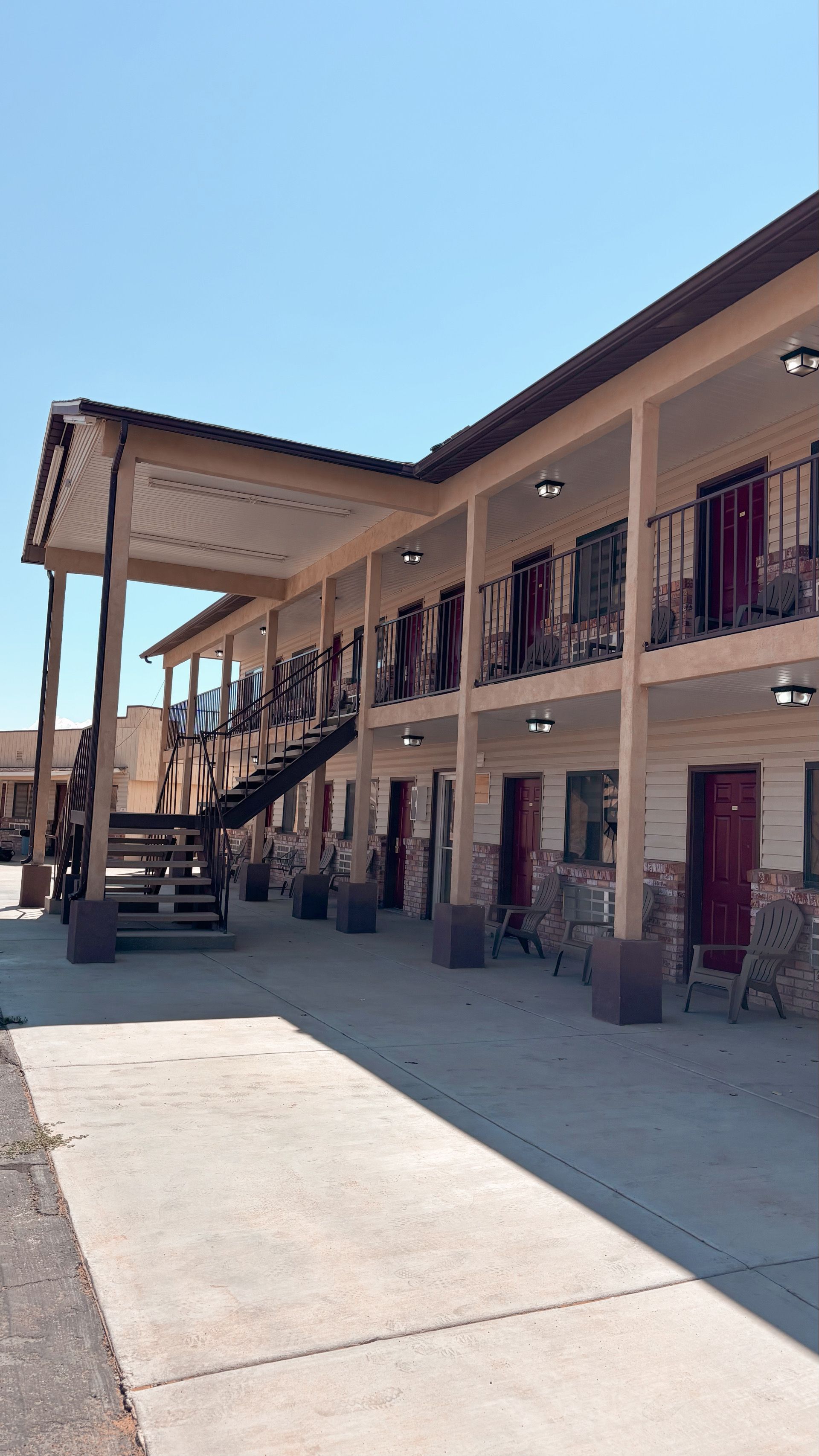 Two-story motel walkway with exterior stairs, brown railings, and open doors under a blue sky.