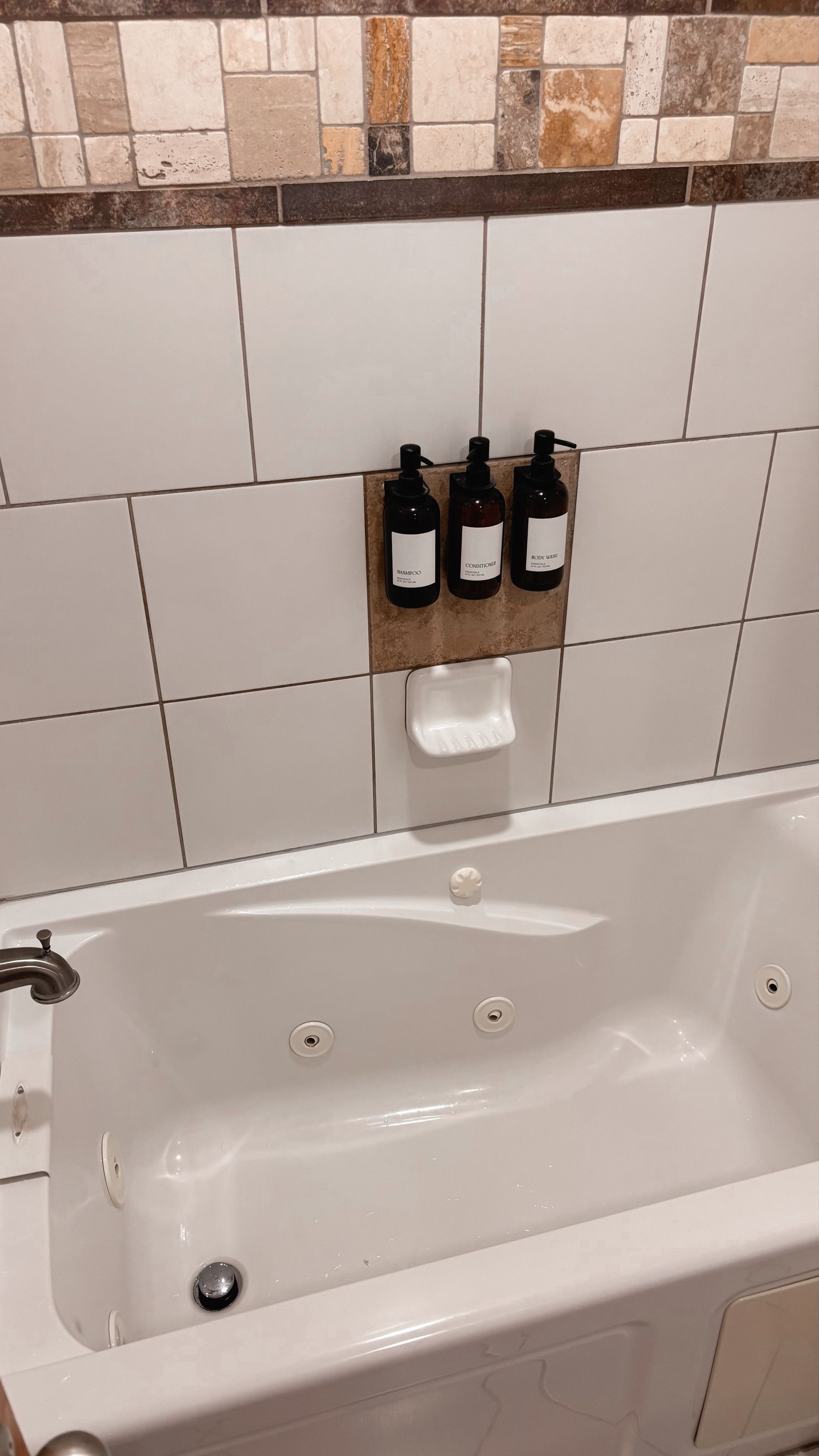 Bathroom tub with white tile wall, decorative border, and three bottles on a shower caddy.