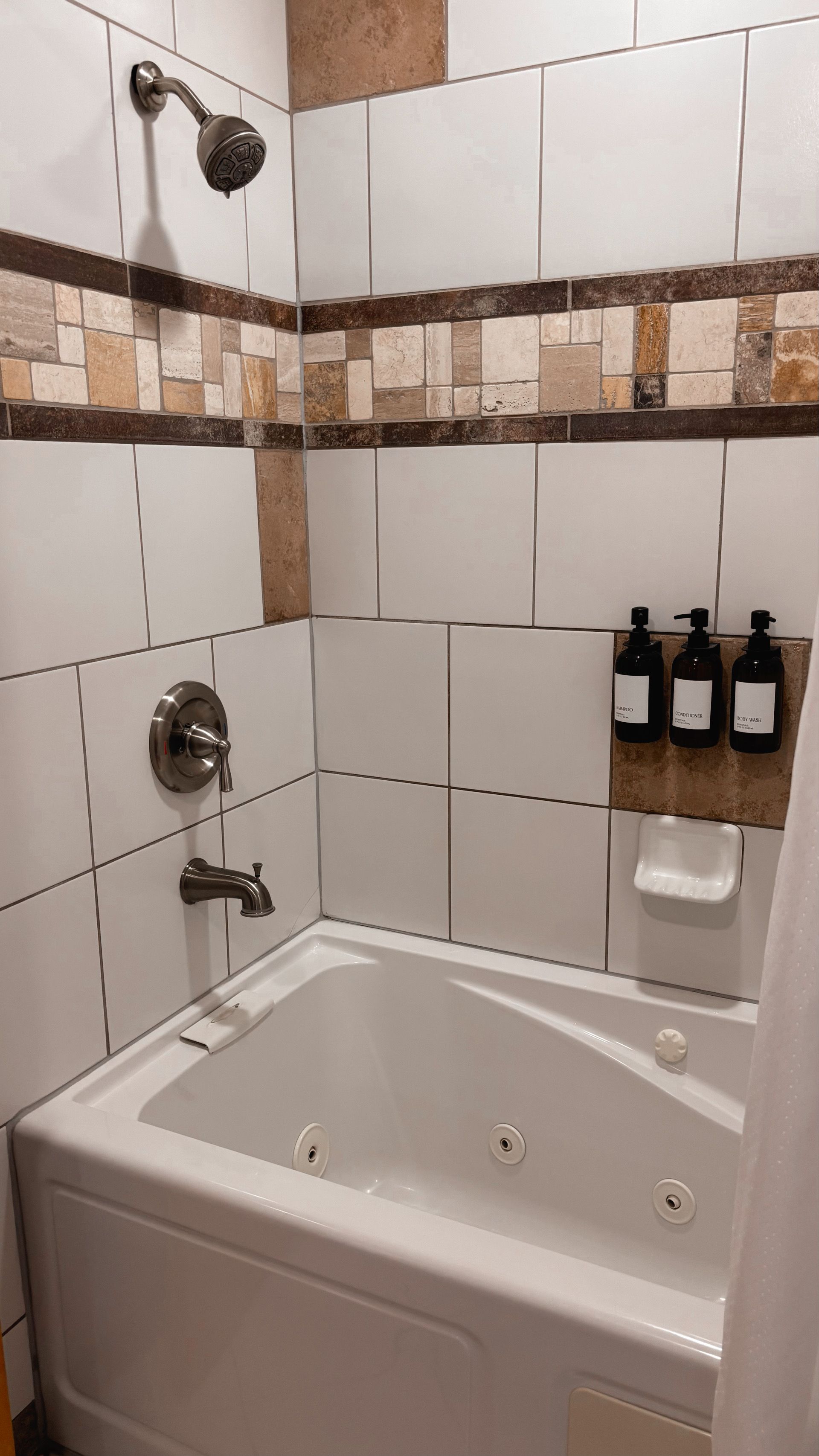 Small white tiled bathroom with a corner soaking tub, shower fixtures, and wall-mounted soap dispensers.