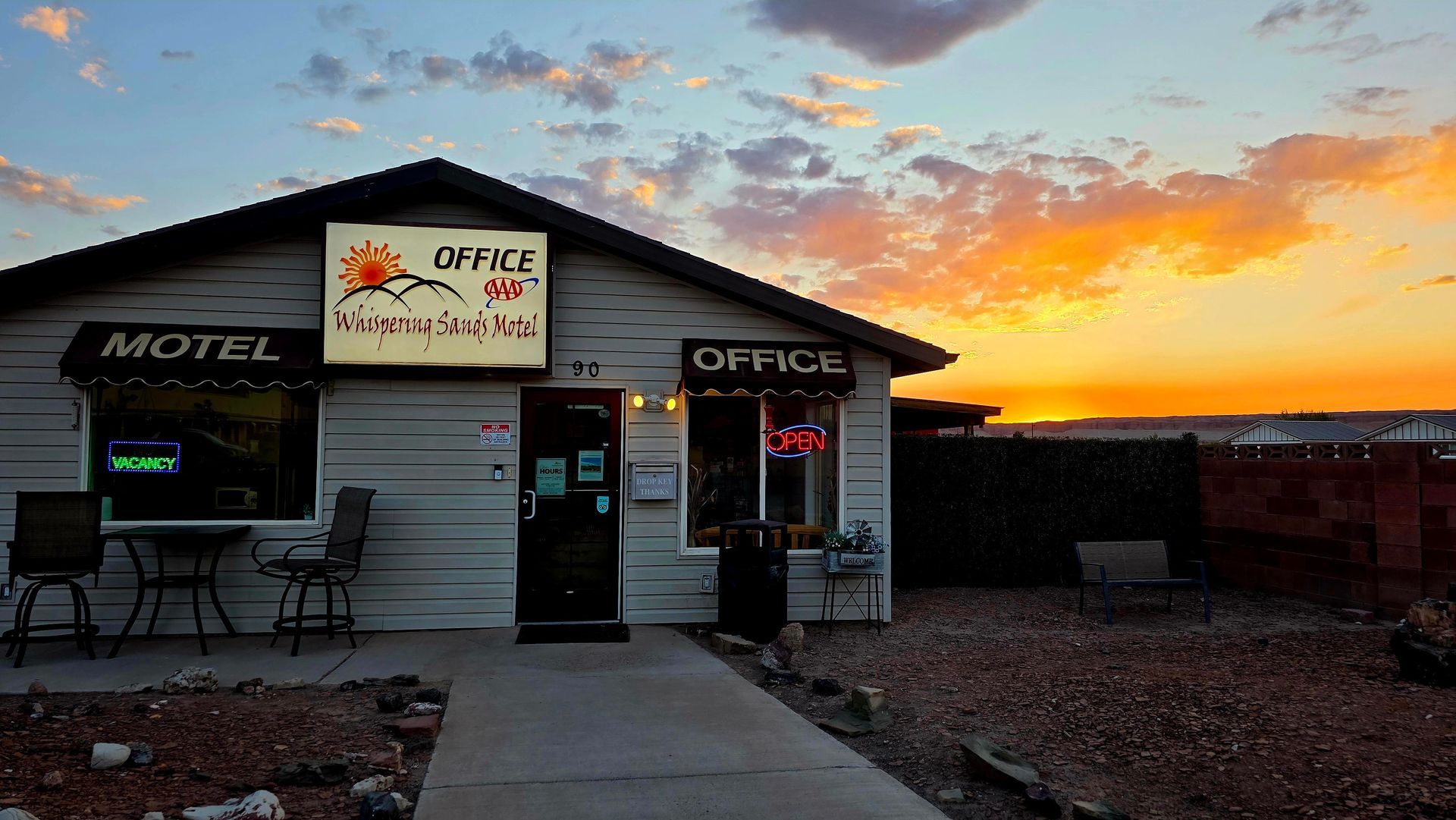 Small roadside motel and office at sunset, with glowing signs, patio seating, and a colorful sky.