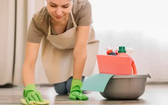 Person in apron and green gloves cleaning a floor beside a bucket and spray bottles