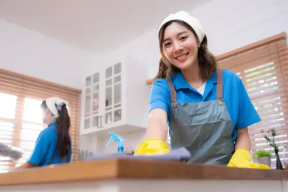 Woman cleaning a kitchen counter with a yellow sponge, wearing a blue shirt and apron.