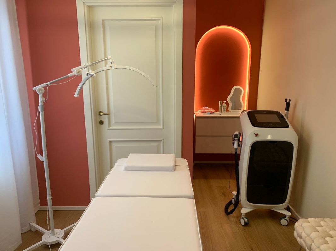 Sala visite mediche con lettino per trattamenti, lampada a soffitto, specchio per il trucco e carrello medico in una nicchia illuminata da luce calda.
