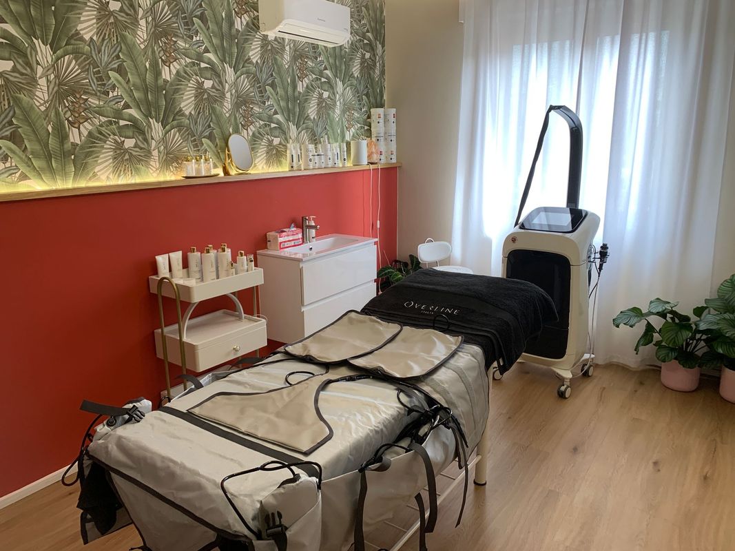 Sala trattamenti in salone con lettino, attrezzature per la bellezza, parete rossa e piante vicino alla finestra.