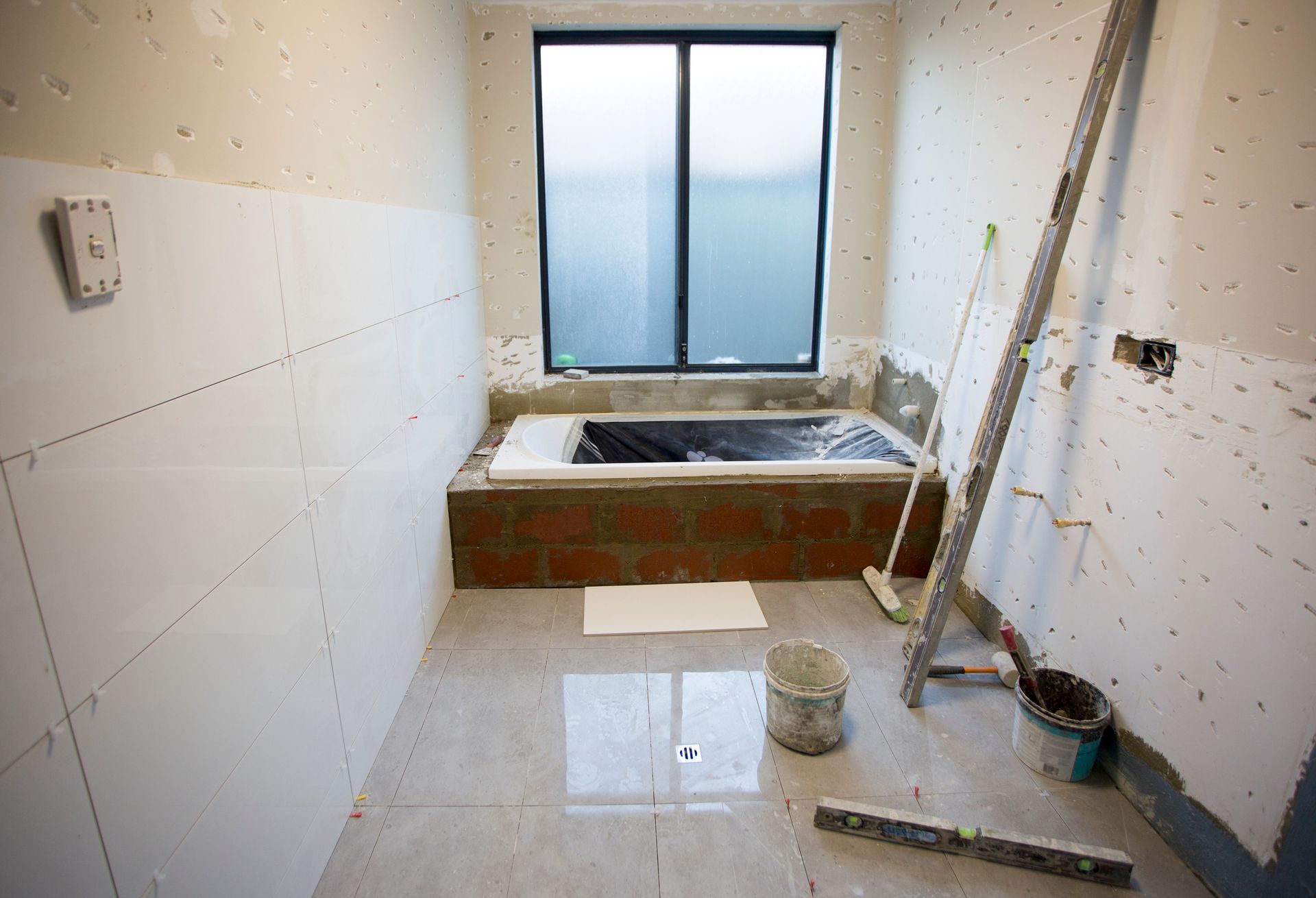 Bathroom under construction with tiled walls and floor, window, and unfinished bathtub. Bathroom under construction with tiled walls and floor, window, and unfinished bathtub.