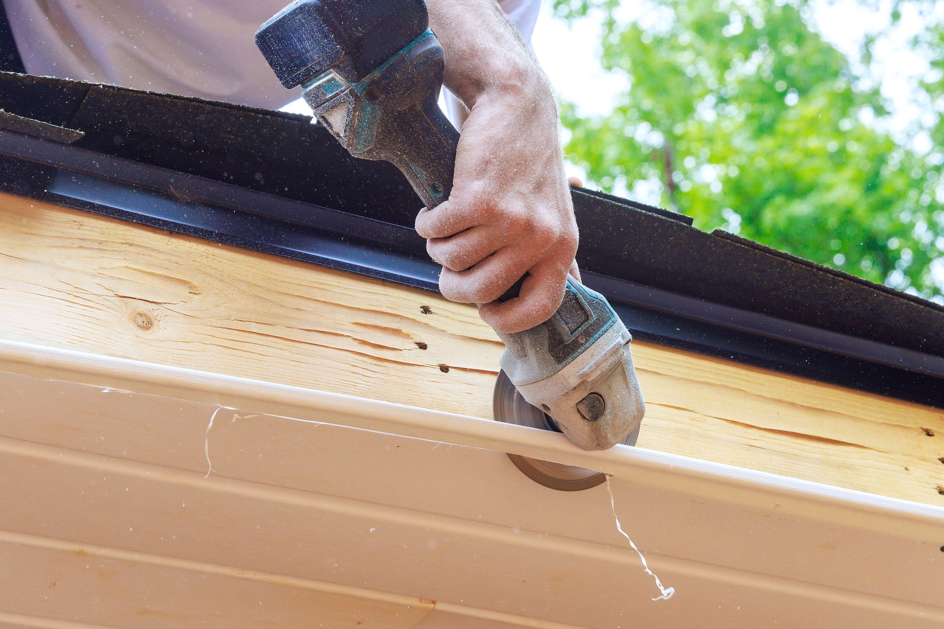 Person uses power tool to cut a white gutter. Wood beams and green foliage in background. Person uses power tool to cut a white gutter. Wood beams and green foliage in background.