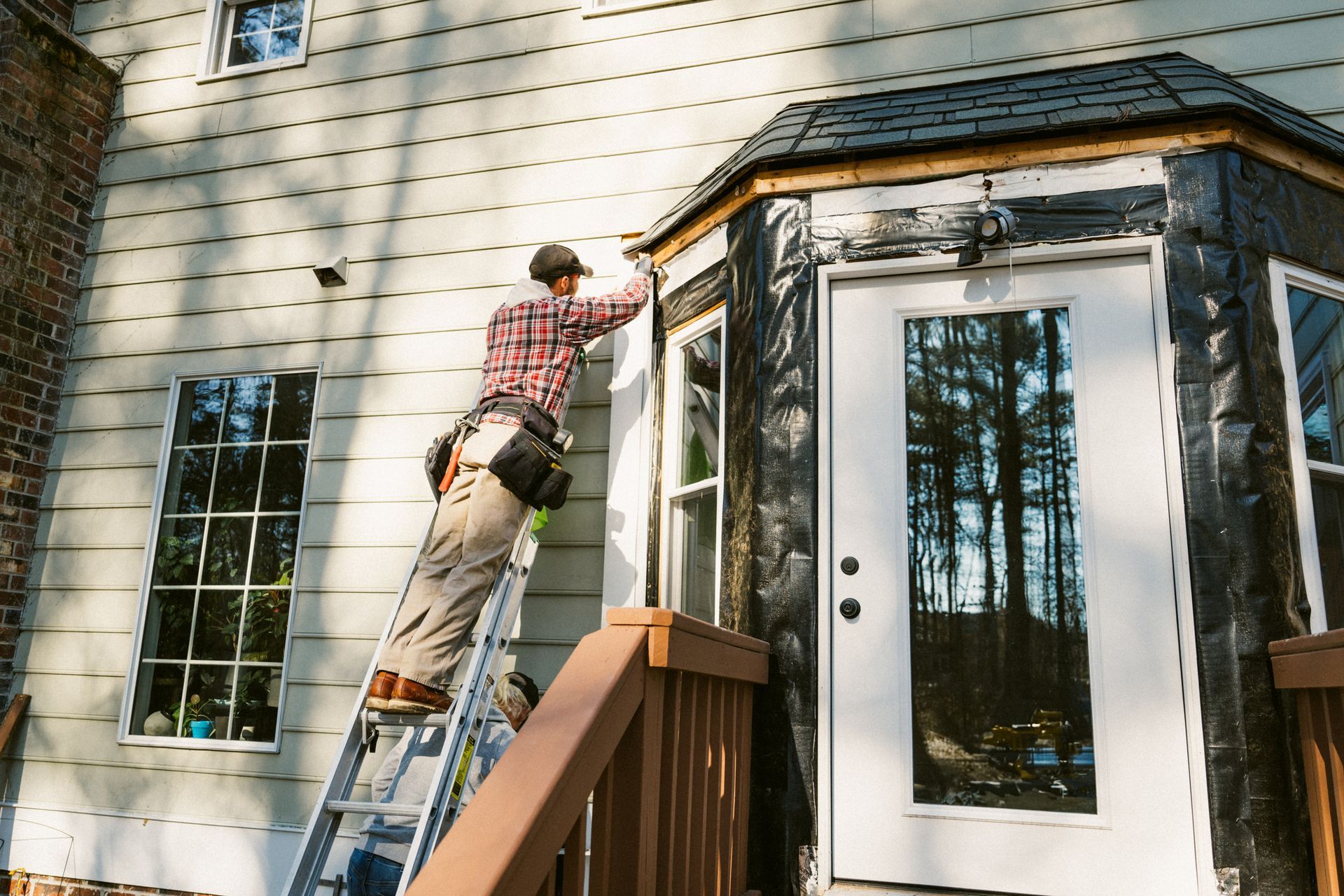 A person on a ladder working on the exterior of a house, near a door and windows. A person on a ladder working on the exterior of a house, near a door and windows.