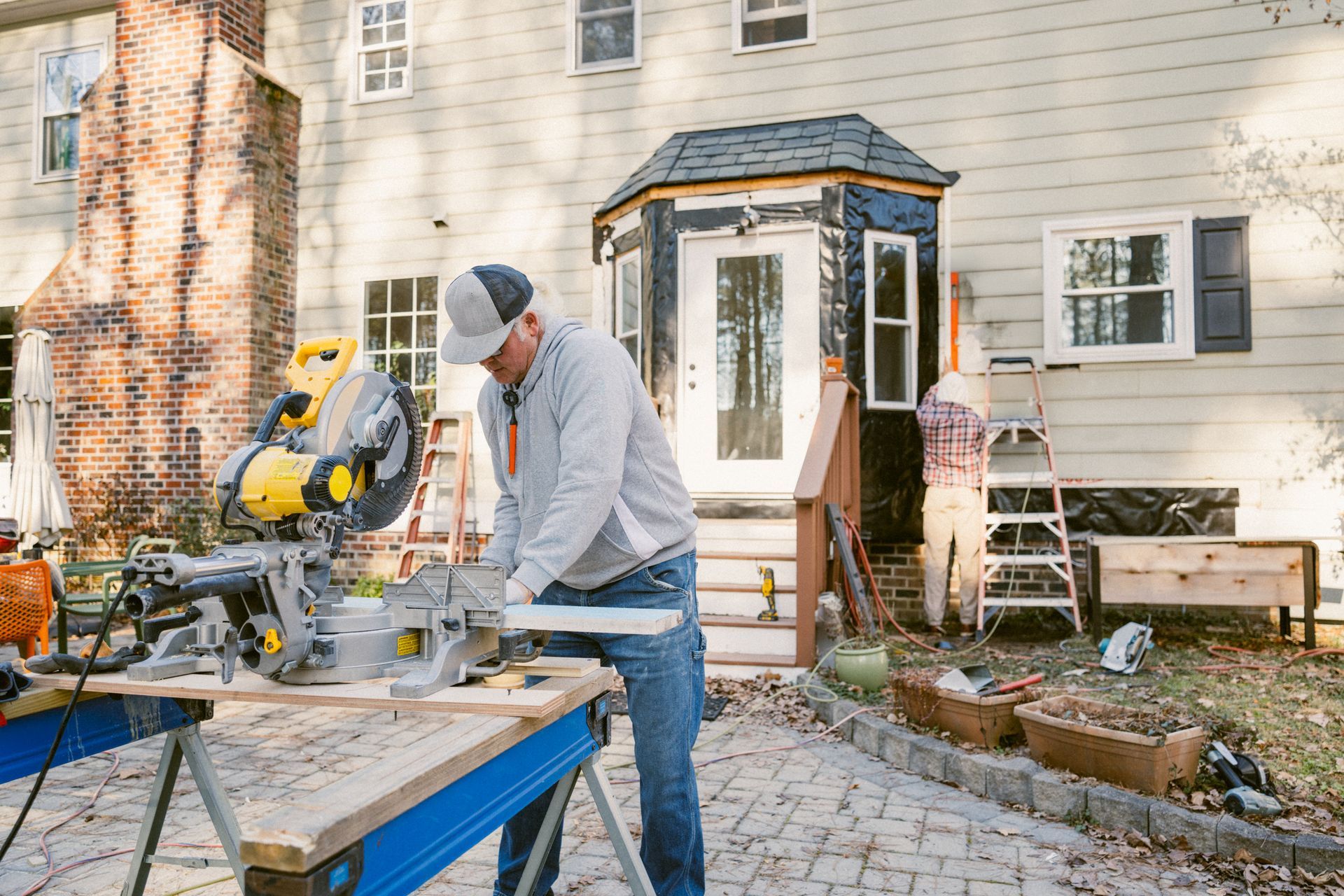 A person using a yellow and black miter saw to cut wood outdoors near a house. A person using a yellow and black miter saw to cut wood outdoors near a house.