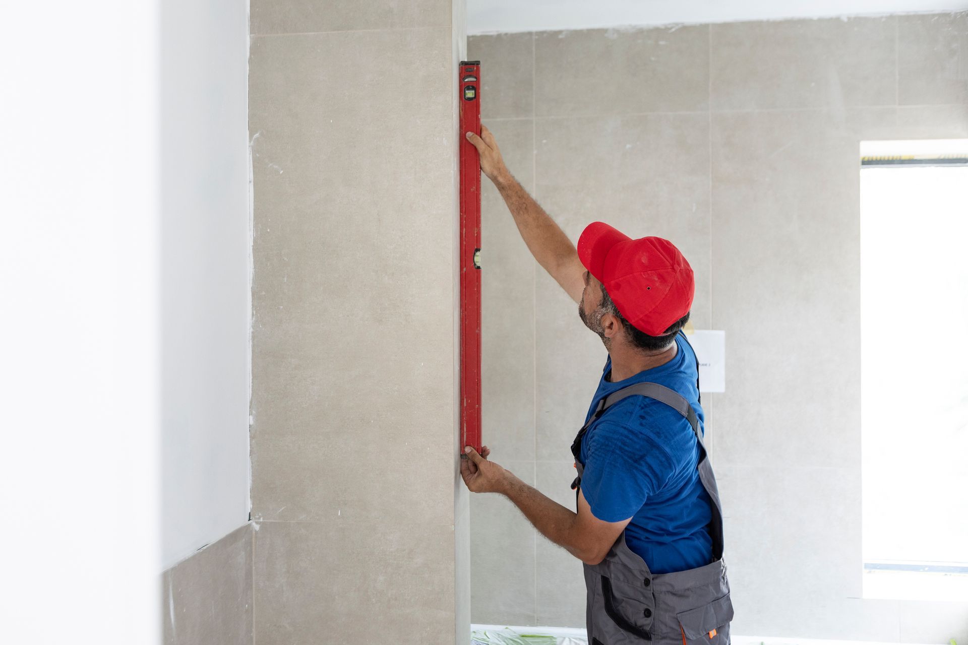 Person in overalls and red cap using a level on a wall in a room. Person in overalls and red cap using a level on a wall in a room.
