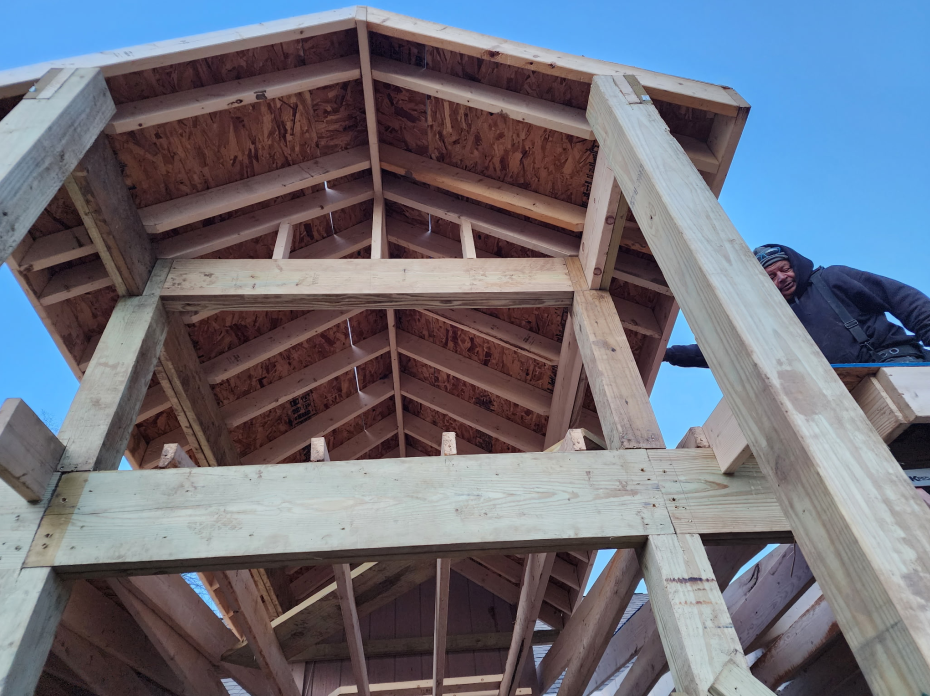 Roofer in overalls and hard hat uses a power drill on a brown metal roof under a blue sky. Roofer in overalls and hard hat uses a power drill on a brown metal roof under a blue sky.