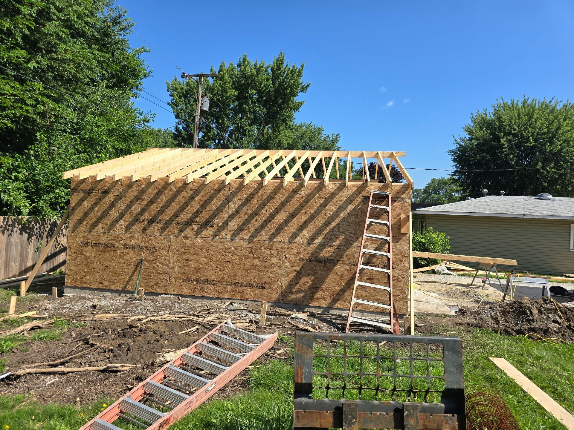 Garage under construction, exterior view: OSB siding, wooden roof supports, ladder, blue sky.