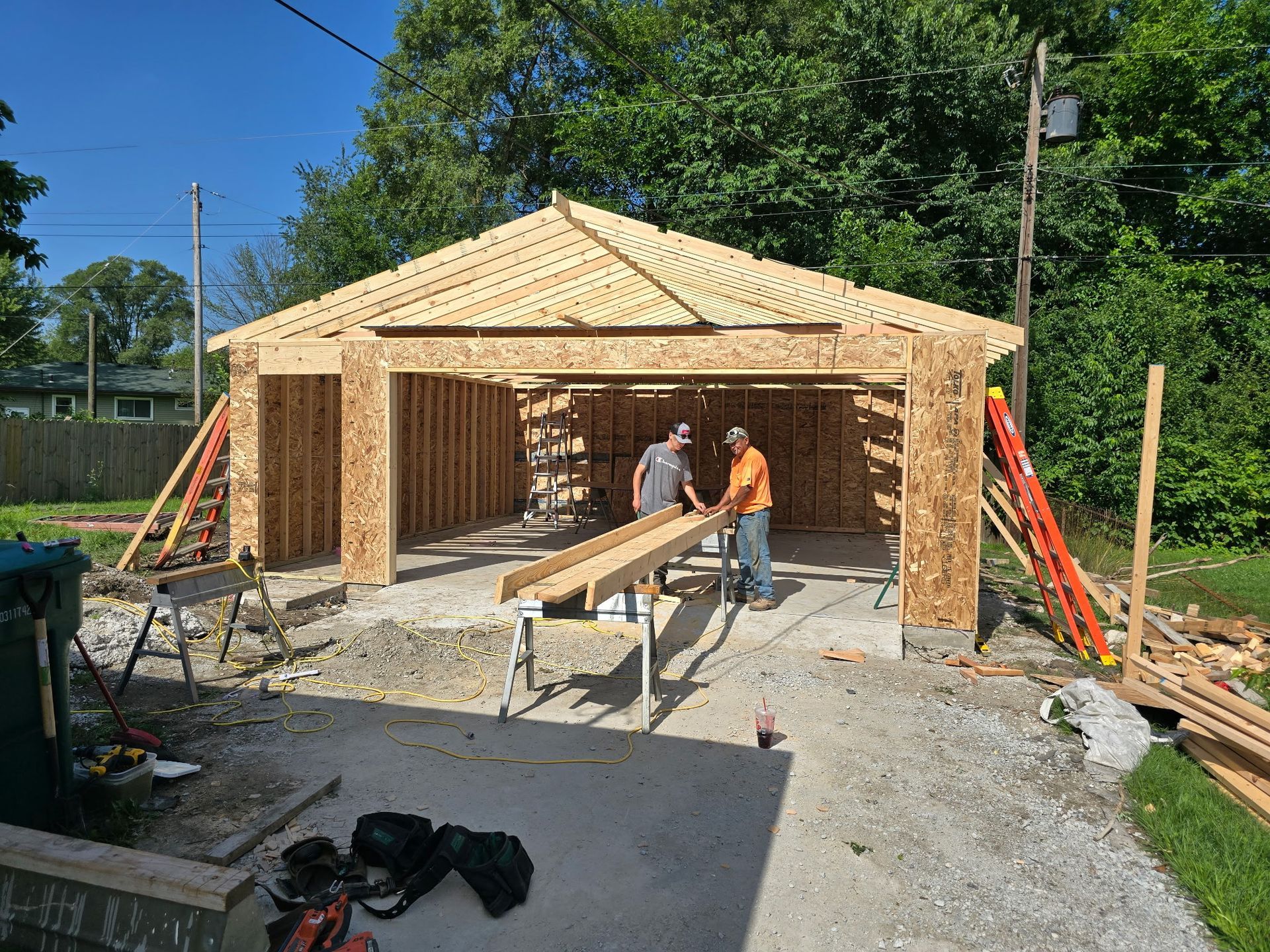 Two people building a wooden garage. One measures a board, others prepare to cut. Sunny day.